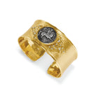 A wide gold cuff ring featuring a central oval dark gray coin with a carved animal motif, accented by small clear stones on a white background.